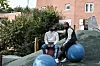 To menn sitter på blå treningsballer ved et grønt uteområde foran en murbygning.