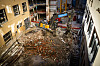 Oversikt over en byggeplass med gravemaskin, stillaser og store mengder rivningsmasser mellom eldre bygninger.