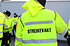 Person i gul refleksjakke med teksten «STREIKEVAKT», omgitt av andre vakter utenfor en bygning.