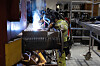 Verkstedarbeider i verneutstyr sveiser på en stor metallkomponent i et togverksted.