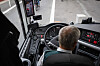 Bussjåfør sett bakfra ved rattet i en moderne buss med instrumentpanel og skjermer.