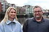 To personer står foran en vannkant og bygninger; kvinnen har blå genser, og mannen bruker briller.