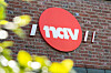 Rød NAV-logo på en teglsteinsvegg, delvis innrammet av uskarpe grønne blader.